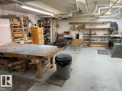 Workshop area featuring a large wooden workbench, exposed overhead piping, concrete flooring, and extensive wall shelving - 