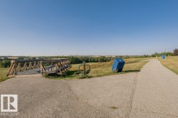 Paved pathway with a wooden pedestrian bridge, surrounded by expansive green space and distant treeline - 