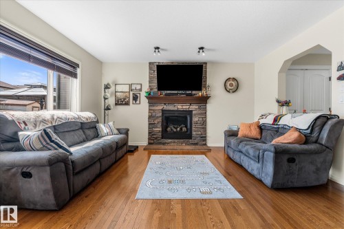 Living room featuring a stone-clad fireplace with a wood mantel, hardwood flooring, and a large window with blinds - 41 Heron Crescent, Spruce Grove, AB - Indoor Photo Showing Living Room With Fireplace