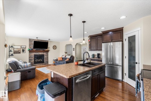 Open-concept kitchen featuring an island with a double basin sink, dark wood cabinetry, stainless steel appliances, and wood-finish flooring - 41 Heron Crescent, Spruce Grove, AB - Indoor Photo Showing Kitchen With Fireplace With Double Sink With Upgraded Kitchen