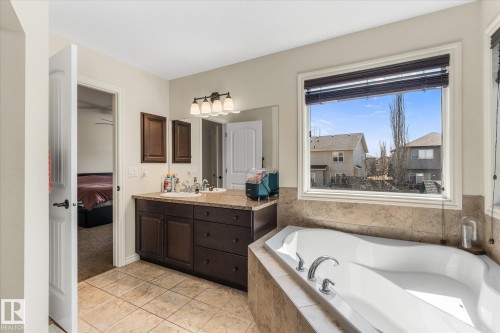 Bathroom featuring a built-in soaking tub with tile surround, a dual vanity with a light-colored countertop, dark wood-finish cabinetry, and a large window with blinds - 41 Heron Crescent, Spruce Grove, AB - Indoor Photo Showing Bathroom