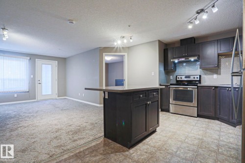 Open-concept kitchen featuring dark cabinetry, stainless steel appliances, and a central island with countertop seating - 201 1520 Hammond Gate Gate, Edmonton, AB - Indoor Photo Showing Kitchen With Stainless Steel Kitchen