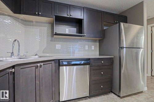Kitchen featuring dark wood-finish cabinetry, light-toned countertops, a stainless steel refrigerator, and a stainless steel dishwasher - 201 1520 Hammond Gate Gate, Edmonton, AB - Indoor Photo Showing Kitchen With Stainless Steel Kitchen With Upgraded Kitchen