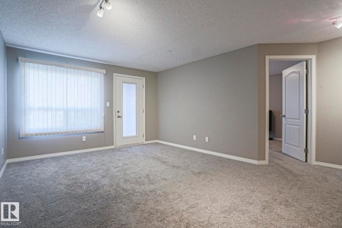 Spacious room featuring neutral paint, gray carpeting, a glass-paneled door, track lighting, and a large window with vertical blinds - 201 1520 Hammond Gate Gate, Edmonton, AB - Indoor Photo Showing Other Room