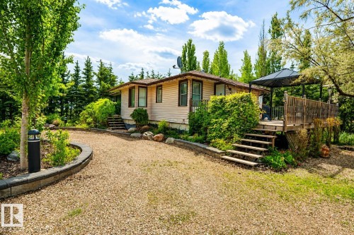 Cream-colored siding residence featuring a gravel driveway, mature trees, and a multi-level wood deck with a dark-colored gazebo - 400 60501 Range Road 120, Rural St. Paul County, AB - Outdoor