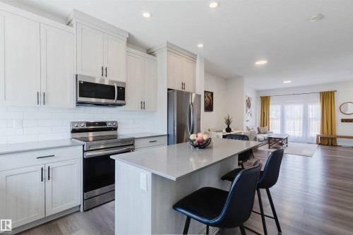 Modern kitchen featuring light wood-finish flooring, white flat-panel cabinetry with black hardware, stainless steel appliances, a white subway tile backsplash, and a light-toned countertop with an integrated breakfast bar - 5237 Edgemont Boulevard, Edmonton, AB - Indoor Photo Showing Kitchen With Stainless Steel Kitchen With Upgraded Kitchen