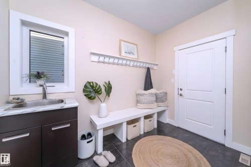 Functional utility room featuring a built-in sink with a white marble-look countertop, dark wood-finish cabinetry, a window with white trim, a coat rack with a shelf, and a white panel door - 8967 24 Avenue, Edmonton, AB - Indoor