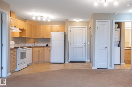 Open concept living space featuring a full kitchen with wood-finish cabinetry and white appliances, a stacked washer and dryer unit, a paneled entry door, and track lighting fixtures - #314 240 Spruce Ridge Rd, Spruce Grove, AB - Indoor Photo Showing Kitchen