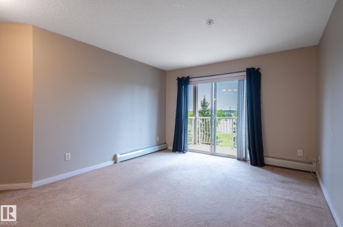 Expansive room featuring neutral wall tones, light-colored carpeting, a sliding glass door, an outdoor balcony with white railings, and baseboard heating - #314 240 Spruce Ridge Rd, Spruce Grove, AB - Indoor Photo Showing Other Room