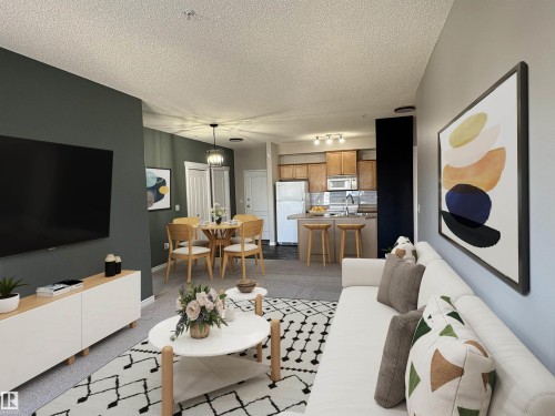 Open-concept living space with carpeted flooring, a dedicated dining area featuring a pendant light fixture, and a kitchen with wood cabinetry and stainless steel appliances - 224 16807 100 Avenue, Edmonton, AB - Indoor Photo Showing Living Room