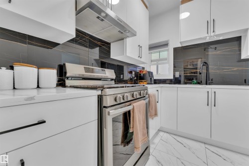Modern kitchen featuring white cabinetry with dark hardware, stainless steel range with overhead ventilation, white countertop, and a black and gold veined backsplash - 1809 18A Avenue, Edmonton, AB - Indoor Photo Showing Kitchen