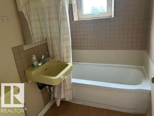 Bathroom featuring a white bathtub, light-toned wall tiling, wood-finish flooring, and a wall-mounted sink with chrome faucet - 2 6920 101 Avenue Nw, Edmonton, AB - Indoor Photo Showing Bathroom