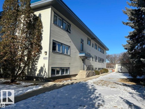 Multi-story building featuring light-colored horizontal siding, multiple windows with white frames, and a prominent address number - 2 6920 101 Avenue Nw, Edmonton, AB - Outdoor