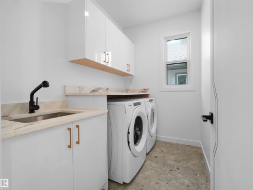Dedicated laundry area featuring a stainless steel sink with matte black faucet, light stone-finish countertops, white cabinetry with gold-tone hardware, and a window - 2070 Collip Crescent, Edmonton, AB - Indoor Photo Showing Laundry Room