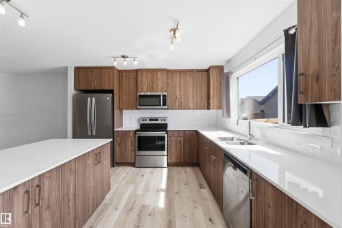 Modern kitchen featuring a large center island with white countertop, wood-finish cabinetry, stainless steel appliances, a double basin sink, and a white tile backsplash - 22371 93 Avenue, Edmonton, AB - Indoor Photo Showing Kitchen With Double Sink With Upgraded Kitchen
