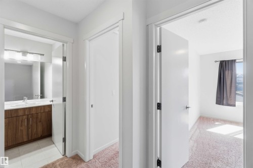 Hallway featuring white trim and doors opening to a bathroom with a wood-finish vanity, light-toned countertop, and mirror, alongside a room with light-toned carpet and a window - 22371 93 Avenue, Edmonton, AB - Indoor