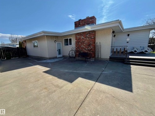 Expansive concrete patio area featuring a brick chimney, a white stucco exterior, and a multi-level wood deck with steps - 16536 103 Street, Edmonton, AB - Outdoor