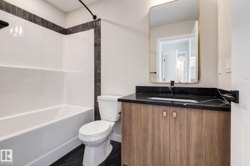 Bathroom featuring a wood-finish vanity with a dark countertop and integrated sink, a rectangular gold-framed mirror, a white toilet, and a white tub with dark tile surround - 44 16231 19 Avenue, Edmonton, AB - Indoor Photo Showing Bathroom