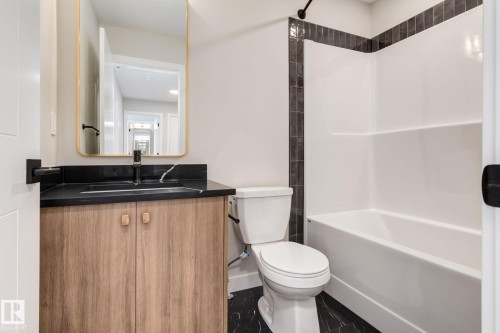Bathroom featuring a wood-finish vanity with a black countertop and integrated sink, a rectangular gold-framed mirror, a white toilet, and a white bathtub with dark tile surround - 44 16231 19 Avenue, Edmonton, AB - Indoor Photo Showing Bathroom