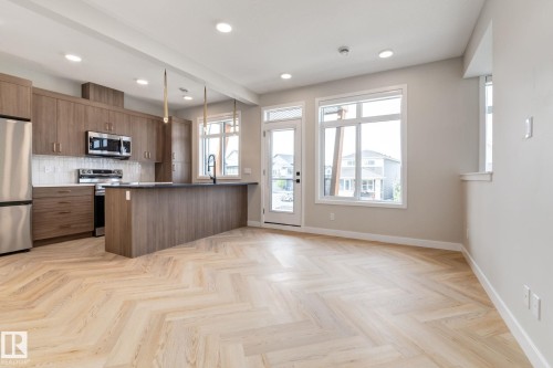 Open-concept living space featuring wood-finish herringbone flooring, a kitchen island with pendant lighting, stainless steel appliances, and large windows providing natural light - 44 16231 19 Avenue, Edmonton, AB - Indoor Photo Showing Kitchen
