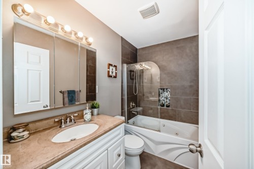 Bathroom featuring a vanity with a light-colored countertop and white cabinetry, a large mirror with overhead lighting, and a bathtub with a glass shower screen and dark tiled surround - 9813 152 Street, Edmonton, AB - Indoor Photo Showing Bathroom