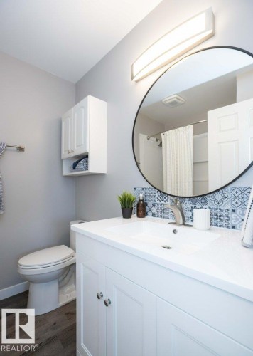 Bathroom featuring a white vanity with integrated sink, patterned tile backsplash, circular wall mirror, and wood-finish flooring - 10812 109 Street, Westlock, AB - Indoor Photo Showing Bathroom