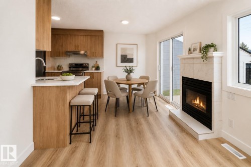 Open-concept living space featuring a tiled fireplace with hearth, wood-finish flooring, and a kitchen with wood cabinetry and light countertops - #63 1033 Youville Dr W Nw, Edmonton, AB - Indoor With Fireplace