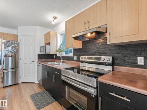The kitchen features stainless steel appliances, wood-look flooring, light wood upper cabinetry, and dark lower cabinetry with a stacked stone-style backsplash - 21360 88 Avenue, Edmonton, AB - Indoor Photo Showing Kitchen With Double Sink