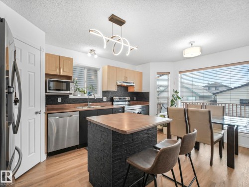 This kitchen features light wood cabinetry, stainless steel appliances, a center island with a stone facade, and a dining area with large windows - 21360 88 Avenue, Edmonton, AB - Indoor