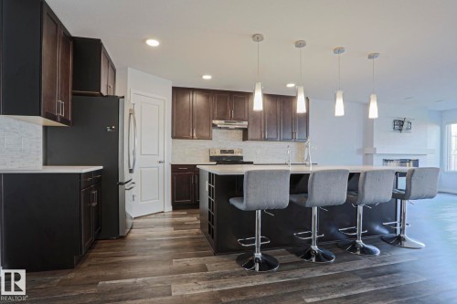 Open-concept kitchen featuring an island with built-in shelving, pendant lighting, wood-finish flooring, dark cabinetry, and a light-toned tile backsplash - 4314 Cooke Lane, Edmonton, AB - Indoor Photo Showing Kitchen With Upgraded Kitchen