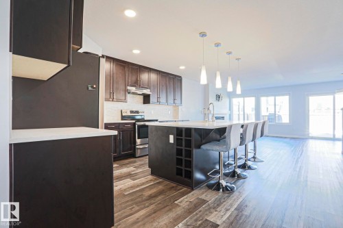 Open-concept kitchen featuring dark wood cabinetry, stainless steel appliances, a white countertop island with integrated wine storage, pendant lighting, and wood-finish flooring - 4314 Cooke Lane, Edmonton, AB - Indoor Photo Showing Kitchen With Stainless Steel Kitchen With Upgraded Kitchen