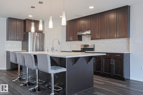 Spacious kitchen featuring an expansive center island with seating, dark wood-finish cabinetry, light-toned stone backsplash, stainless steel appliances, and dark wood-finish flooring - 4314 Cooke Lane, Edmonton, AB - Indoor Photo Showing Kitchen With Stainless Steel Kitchen With Upgraded Kitchen