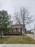 Brown siding exterior with white window frames, a gray shingle roof, mature front yard trees, and a concrete driveway - 