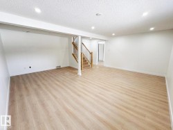 Spacious room featuring light wood-finish flooring, recessed ceiling lighting, and a prominent wood staircase with white risers and railings - 