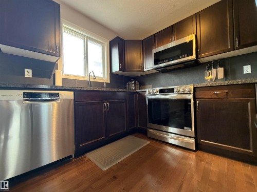 Kitchen featuring rich wood cabinetry, stainless steel appliances, dark granite countertops, a tiled backsplash, and wood-finish flooring - 5204 4 Avenue, Edmonton, AB - Indoor Photo Showing Kitchen