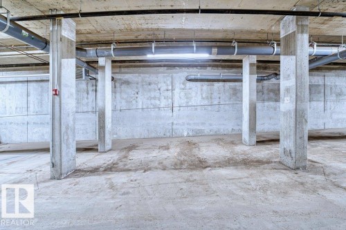 Concrete parking structure featuring exposed overhead piping, robust support columns, and overhead lighting - 318 1316 Windermere Way, Edmonton, AB - Indoor Photo Showing Garage