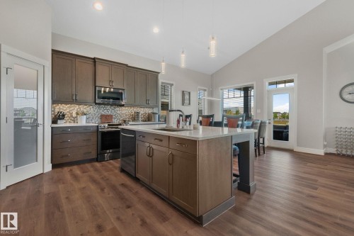 Spacious kitchen featuring wood-finish flooring, an expansive island with a sink, quartz countertops, stainless steel appliances, and a mosaic tile backsplash - 67 Enchanted Way Nw, St. Albert, AB - Indoor Photo Showing Kitchen