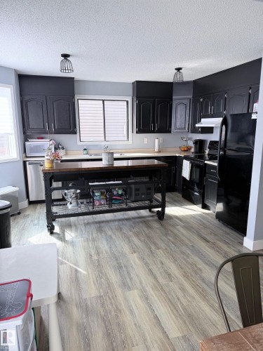 Spacious kitchen featuring extensive black cabinetry, a large window with white blinds, wood-finish flooring, and two modern cage-style pendant lights - 5108 58 Avenue, Elk Point, AB - Indoor Photo Showing Other Room
