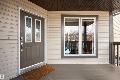 Inviting front entry featuring a brown paneled door with decorative glass inserts, light beige horizontal siding, a white-framed window with grilles, and a wood-finish porch floor - 1205 Ormsby Lane, Edmonton, AB - Outdoor With Deck Patio Veranda With Exterior