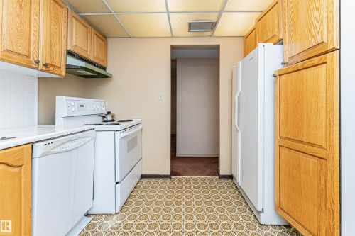 Kitchen featuring wood cabinetry, white appliances, patterned vinyl flooring, and a drop ceiling with recessed lighting - 407 10045 118 Street, Edmonton, AB - Indoor Photo Showing Kitchen