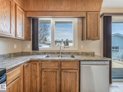 Kitchen featuring a double basin stainless steel sink, wood-finish cabinetry, a white dishwasher, and a window with vertical blinds - 1027 77 Street, Edmonton, AB - Indoor Photo Showing Kitchen With Double Sink