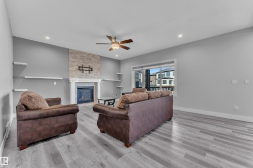 Spacious living area featuring light wood-finish flooring, a stone-accented fireplace with a white mantel, recessed ceiling lights, a ceiling fan, and a large window - 4403 Triomphe Gate, Beaumont, AB - Indoor Photo Showing Living Room With Fireplace