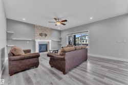 Spacious living area featuring light wood-finish flooring, a stone-accented fireplace with a white mantel, recessed ceiling lights, a ceiling fan, and a large window - 