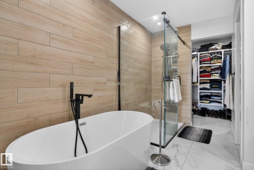 Modern bathroom featuring a freestanding soaking tub with matte black fixtures, a walk-in shower with a clear glass enclosure, wood-finish tile accent wall, and white marble-look floor tiles - 3605 42 Avenue, Beaumont, AB - Indoor Photo Showing Bathroom