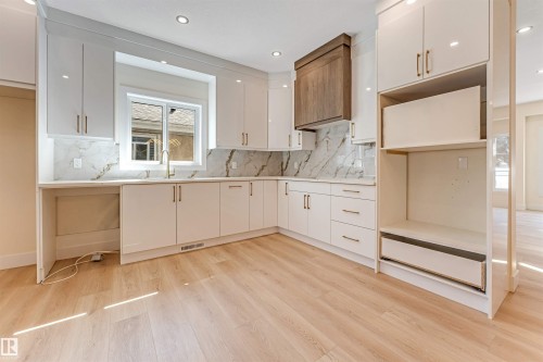 Contemporary kitchen featuring white cabinetry with brushed gold hardware, a large window above the sink, a wood-finish range hood, white countertops, and a marble-look backsplash - 10323 79 Street, Edmonton, AB - Indoor Photo Showing Kitchen