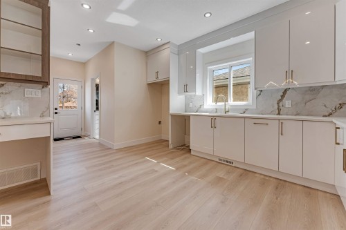 Light wood-finish flooring complements the contemporary kitchen featuring white cabinetry with gold-toned hardware, a sink with a gooseneck faucet, and marble-patterned backsplash and countertops - 10323 79 Street, Edmonton, AB - Indoor Photo Showing Kitchen