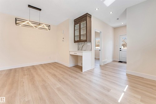 Open-concept living area featuring light wood-finish flooring, a modern linear chandelier, recessed lighting, a built-in cabinet with glass doors and a workstation, and a white paneled door - 10323 79 Street, Edmonton, AB - Indoor Photo Showing Other Room