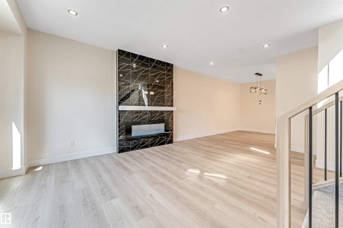 Spacious interior featuring a contemporary fireplace with a dark marble-patterned surround, light wood-finish flooring, recessed lighting, a modern chandelier, and an open staircase with metal balusters - 10323 79 Street, Edmonton, AB - Indoor Photo Showing Other Room With Fireplace