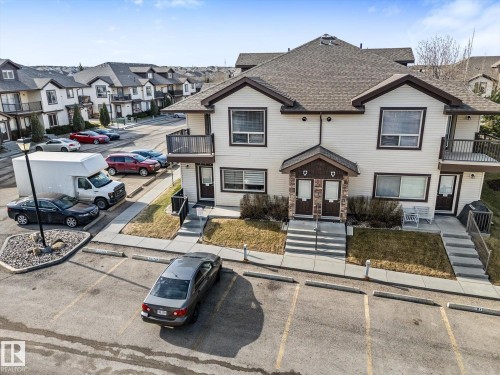 Multi-unit facade featuring light-colored siding with dark trim, a covered entry with stone accents, a second-story balcony with a dark railing, and multiple windows with blinds - 86 604 62 Street, Edmonton, AB - Outdoor