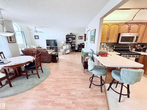 Open-concept living space featuring wood-finish flooring, a kitchen with wood cabinetry and stainless steel appliances, a breakfast bar, and a dining area with a contemporary chandelier - 410 9926 100 Avenue, Fort Saskatchewan, AB - Indoor Photo Showing Dining Room